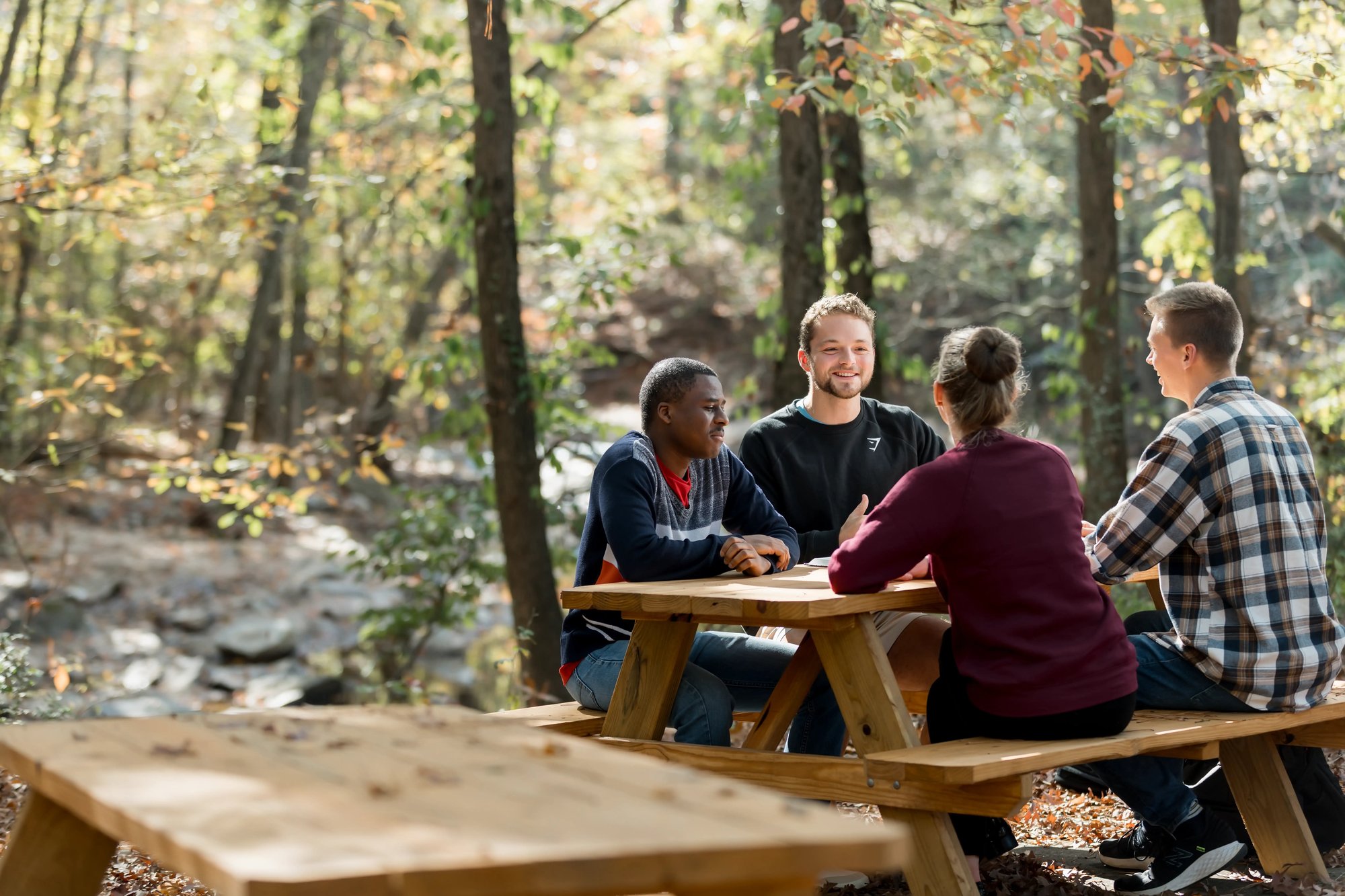 Students sitting at a picnic table outdoors on a wooded campus.