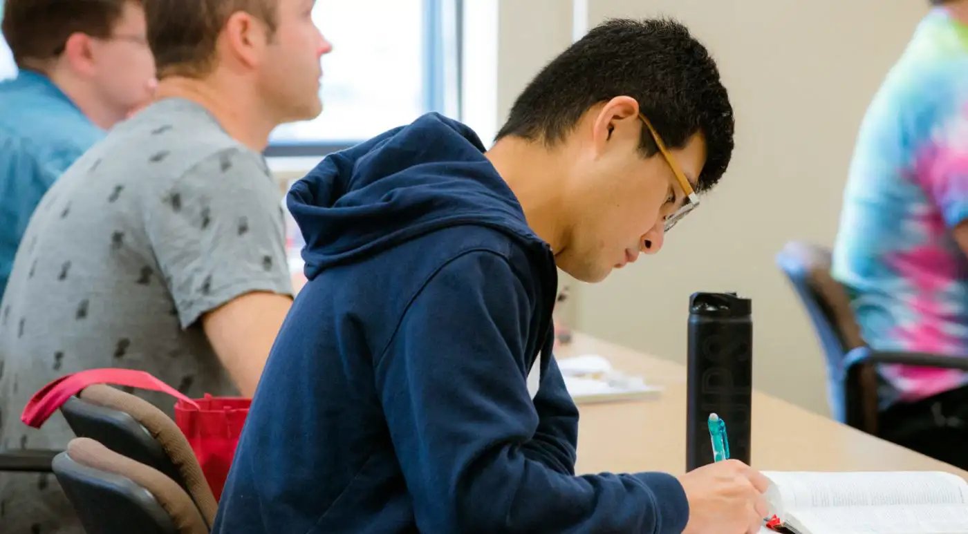 Student taking notes at a classroom desk while other students sit nearby in the background
