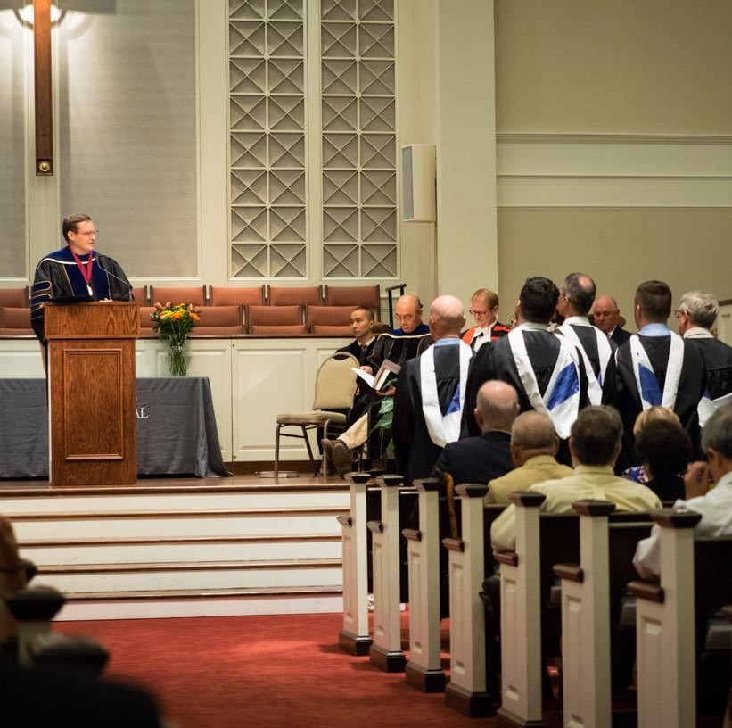 Faculty and graduates gathered during an ordination or commissioning ceremony inside a seminary chapel with a speaker at the pulpit
