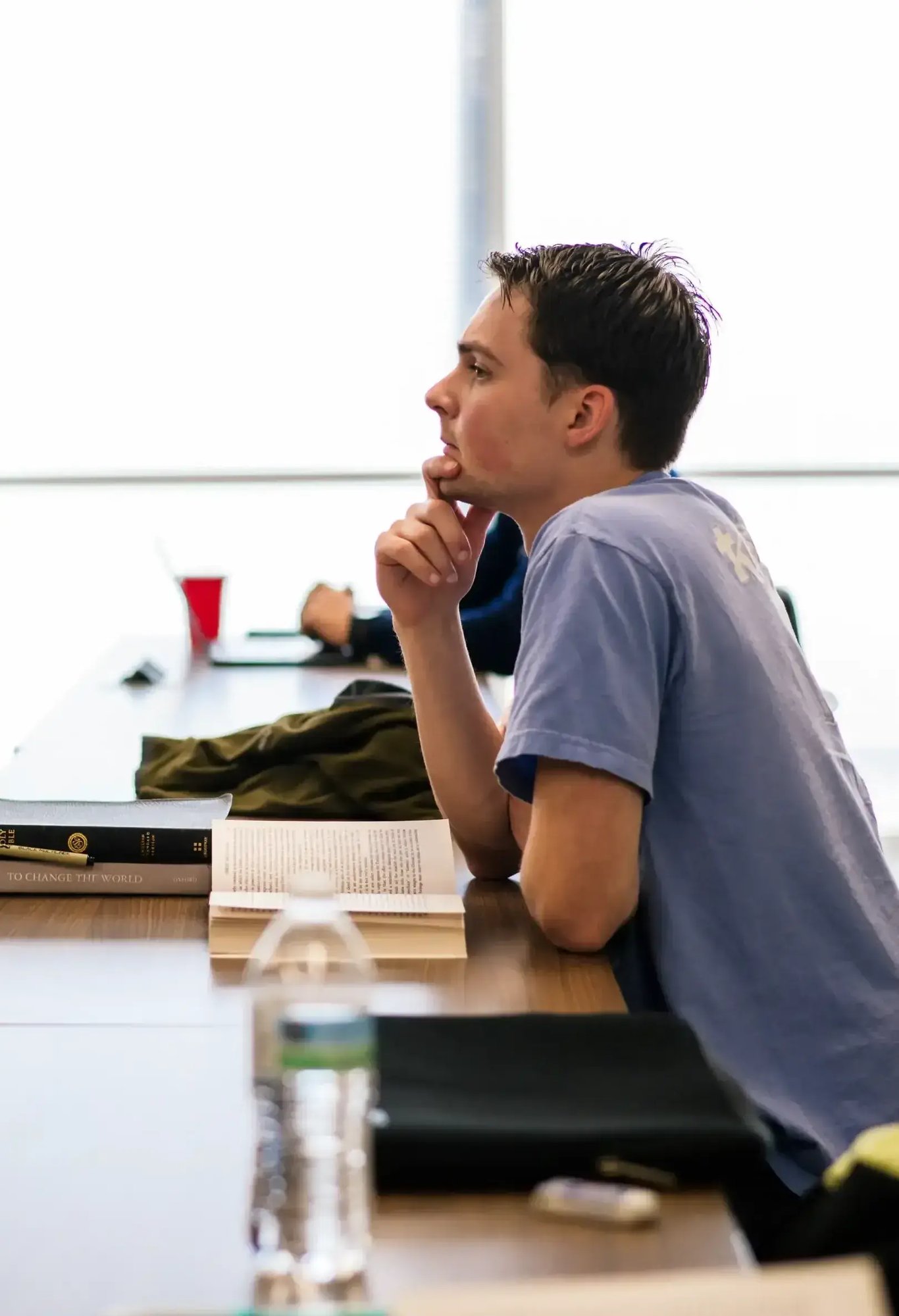 Student seated at a table listening thoughtfully during a classroom Bible study with an open book in front of him