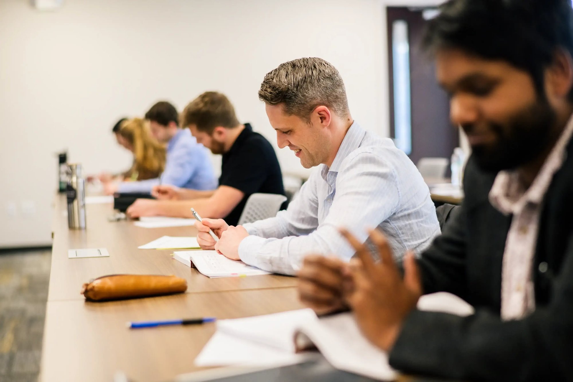 Students seated at a long table taking notes during a classroom lecture in a seminary setting