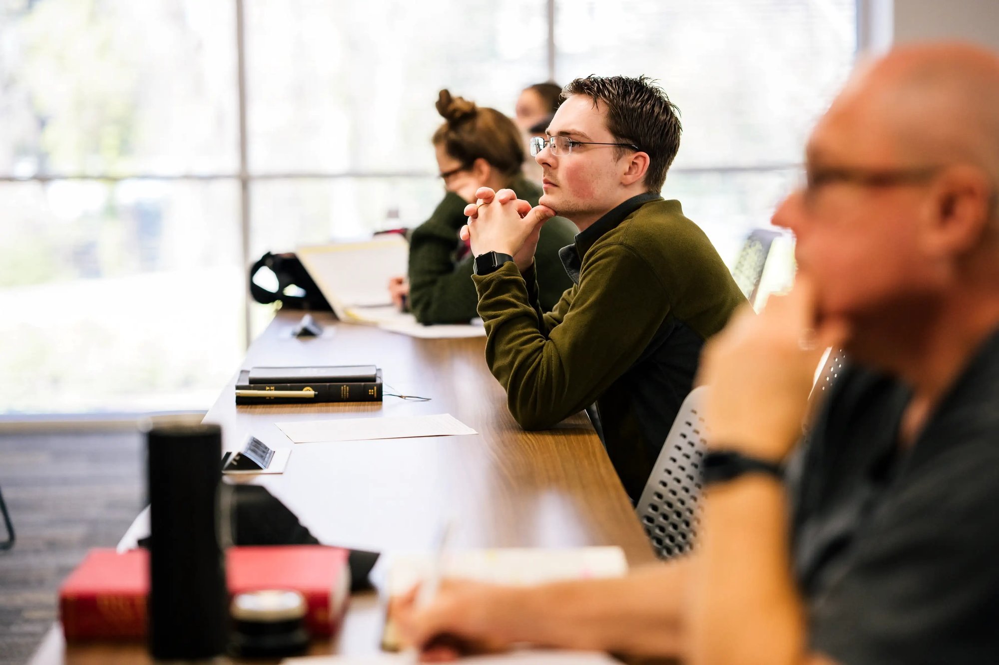 Seminary students seated at a long table listening attentively during a classroom lecture with books and notebooks on the table